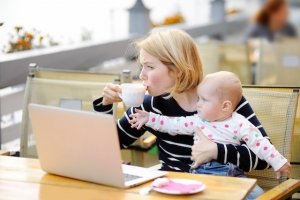 Tired young single mom working oh her laptop, holding daughter and drinking coffee